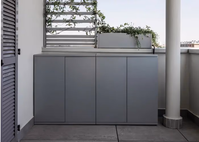 Mobile contenitore grigio a quattro ante installato sul balcone di un terrazzo sotto una fioriera.