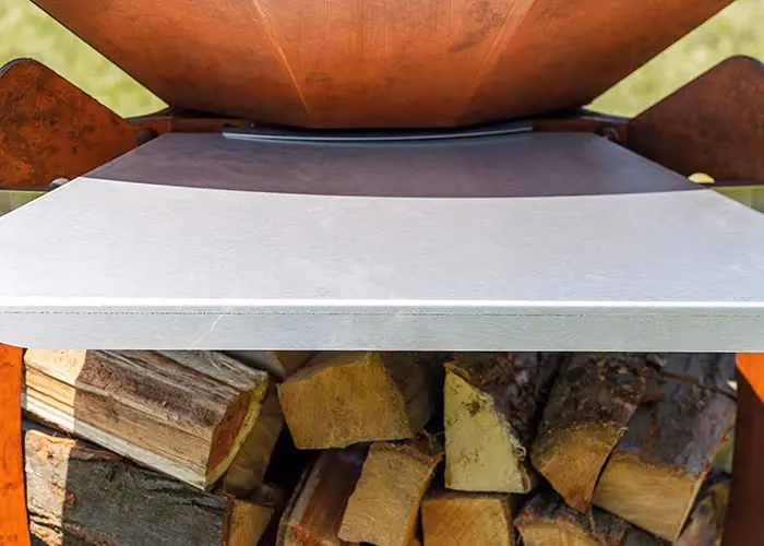 Nahaufnahme der seitlichen Stahlablage eines Grills in Corten-Optik, mit Brennholz im Fach darunter.