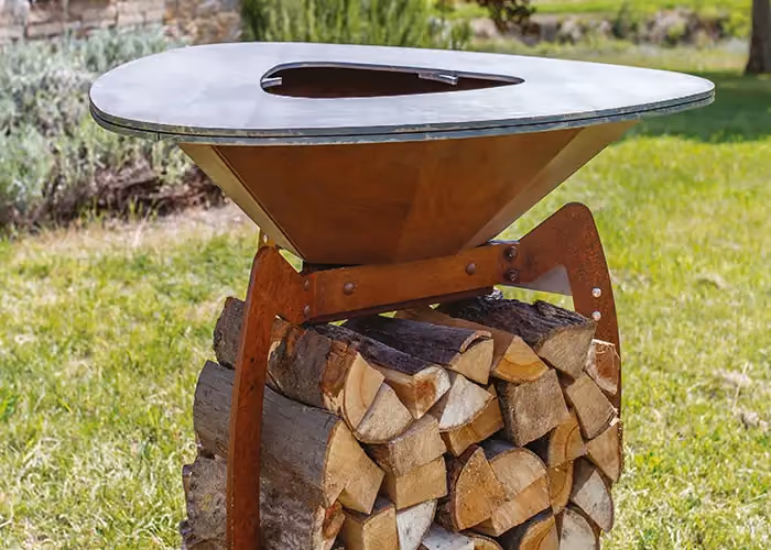 Außengrill in Corten-Optik mit runder Platte und mit Brennholz gefülltem unterem Fach, in einem Garten.