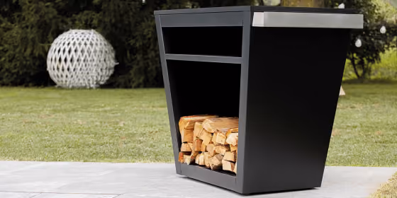 Banco barbecue moderno da esterno in metallo nero con vano inferiore porta legna e piano laterale in acciaio, posizionato in un giardino.