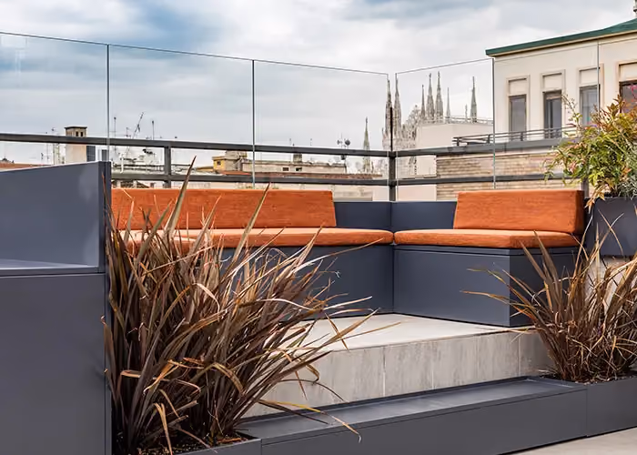 Moderner Loungebereich auf einer Stadtterrasse mit modularen Außensitzmöbeln aus dunklem Metall, orangefarbenen Kissen und integrierten Pflanzgefäßen mit Zierbepflanzung.