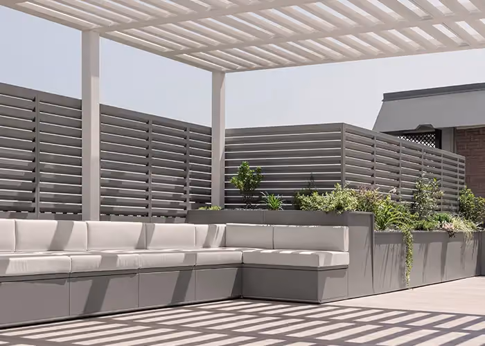 Moderne, von einer Pergola überdachte Terrasse mit langer modularer Außensitzbank mit hellen Kissen und integrierten Pflanzgefäßen.