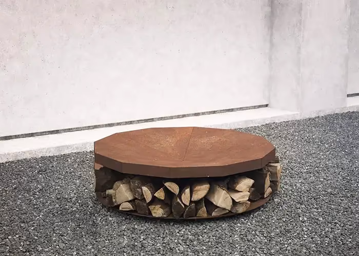 Modern outdoor fire pit with an octagonal corten-effect top and an open bay below holding firewood, standing on grey gravel.