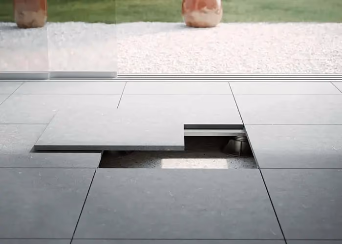 Modern outdoor flooring in modular grey tiles with an access panel lifted, in front of a glazed wall.