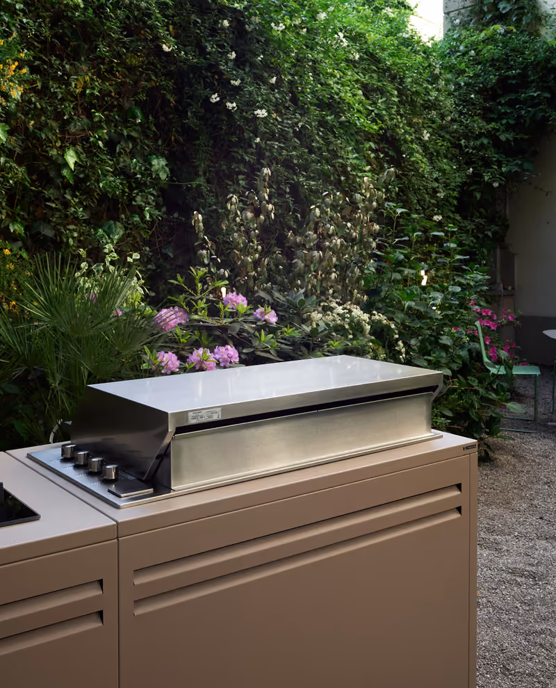 Primo piano del barbecue in acciaio inox integrato nella cucina da esterno beige, circondato da fiori viola.