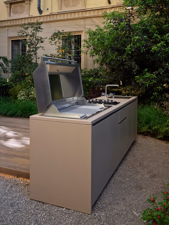 Isola cucina da esterno color tortora con coperchio del barbecue in acciaio aperto, piano cottura e lavello, su ghiaia davanti a un edificio storico