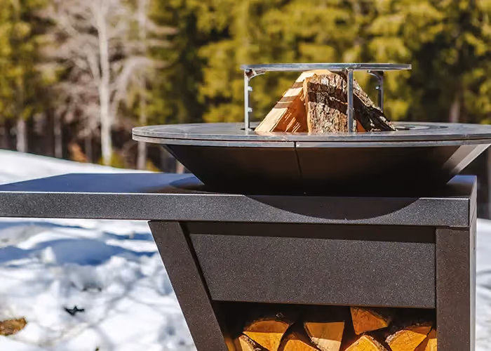Barbecue moderno da esterno con braciere circolare, supporto per legna e vano inferiore porta legna, ambientato in un paesaggio montano innevato.