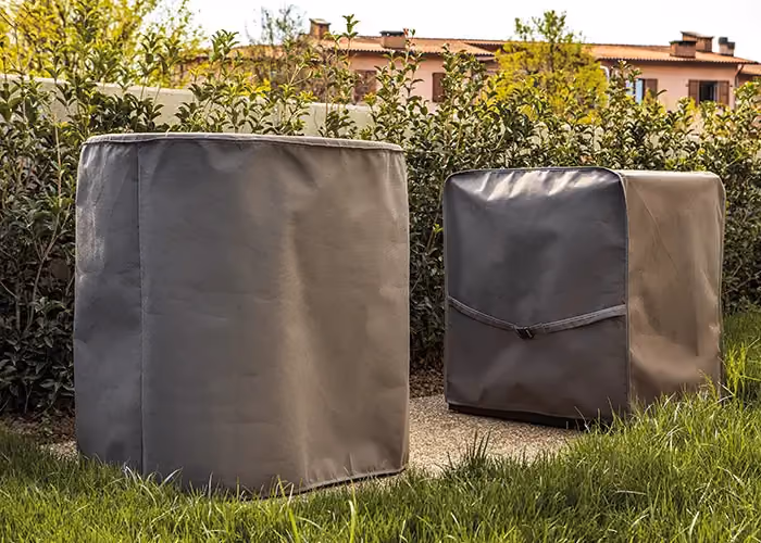 Coperture protettive grigie per barbecue da esterno, posizionate su prato vicino a una siepe.