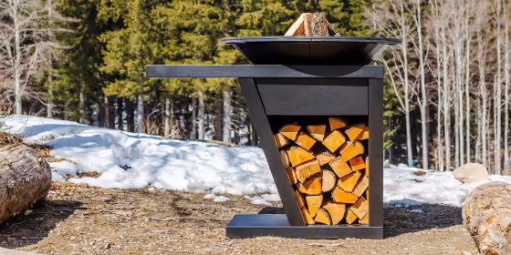 Braciere verticale nero con focolare superiore e vano portalegna acceso, in un bosco innevato