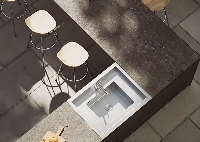 Draufsicht auf eine moderne Außenküche mit Arbeitsplatte in Steinoptik, integrierter Spüle und hellen Hockern auf Natursteinbelag.