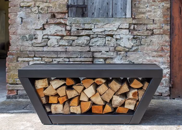Portalegna moderno da esterno con struttura geometrica in metallo scuro e vano centrale riempito con legna da ardere, davanti a un muro rustico in pietra e mattoni.