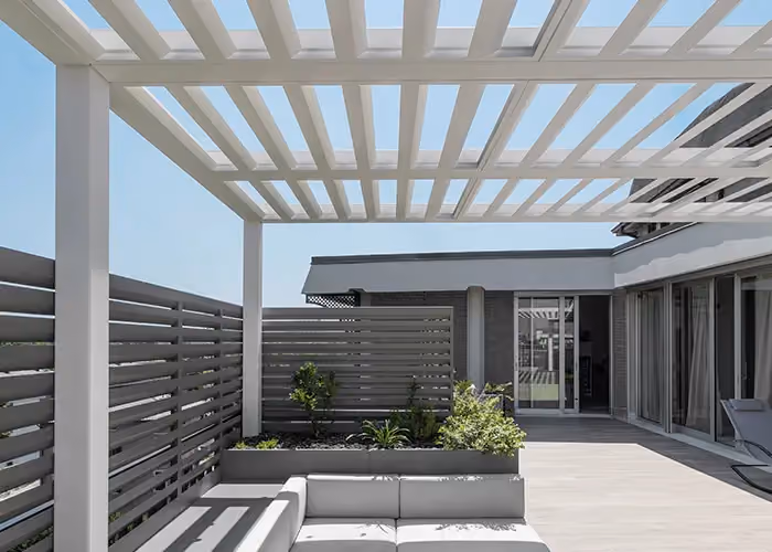 Large modern terrace with a white bioclimatic pergola, grey planters with an integrated sofa, and a glazed façade.