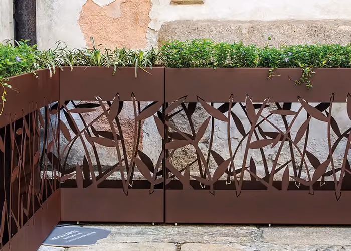 Modulare Pflanzgefäße in Corten-Optik mit dekorativen Ausschnittpaneelen und Zierpflanzen, in einem Außenhof.
