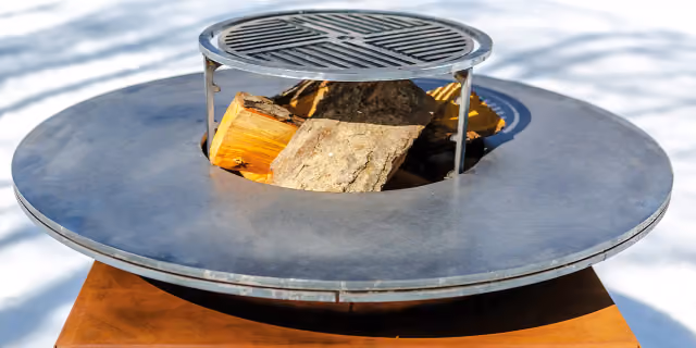 Runde Feuerschale aus Cortenstahl mit mittigem Grillrost und brennendem Feuer, am Pool