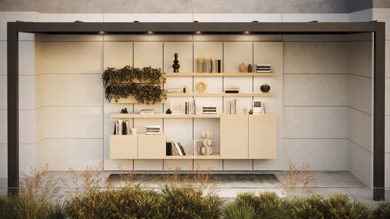 Frontalansicht des Vista-Wall-Systems unter einer dunklen Pergola: offene Regalböden mit Büchern und Objekten, helle Unterschränke und ein Hängepflanzgefäß mit Efeu, vor einer hellen Natursteinwand