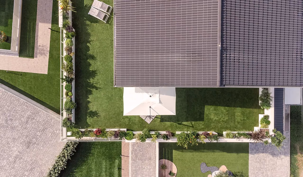 Overhead aerial view of an L-shaped roof garden with artificial turf, a white parasol and a continuous run of flowering perimeter planters along the railing.