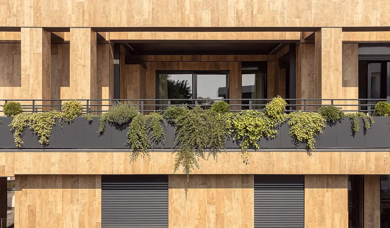 Frontalansicht eines Balkons mit linearen dunklen Pflanzgefäßen voller üppiger grüner Pflanzen, vor einem schwarzen Metallhandlauf an Wänden in Holzoptik.