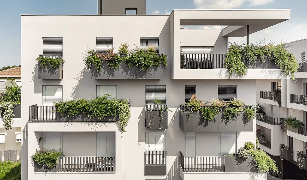 Façade d'un bâtiment blanc aux fenêtres à volets roulants et aux balcons géométriques à garde-corps noirs et bacs rectangulaires gris foncé remplis de plantes grimpantes.