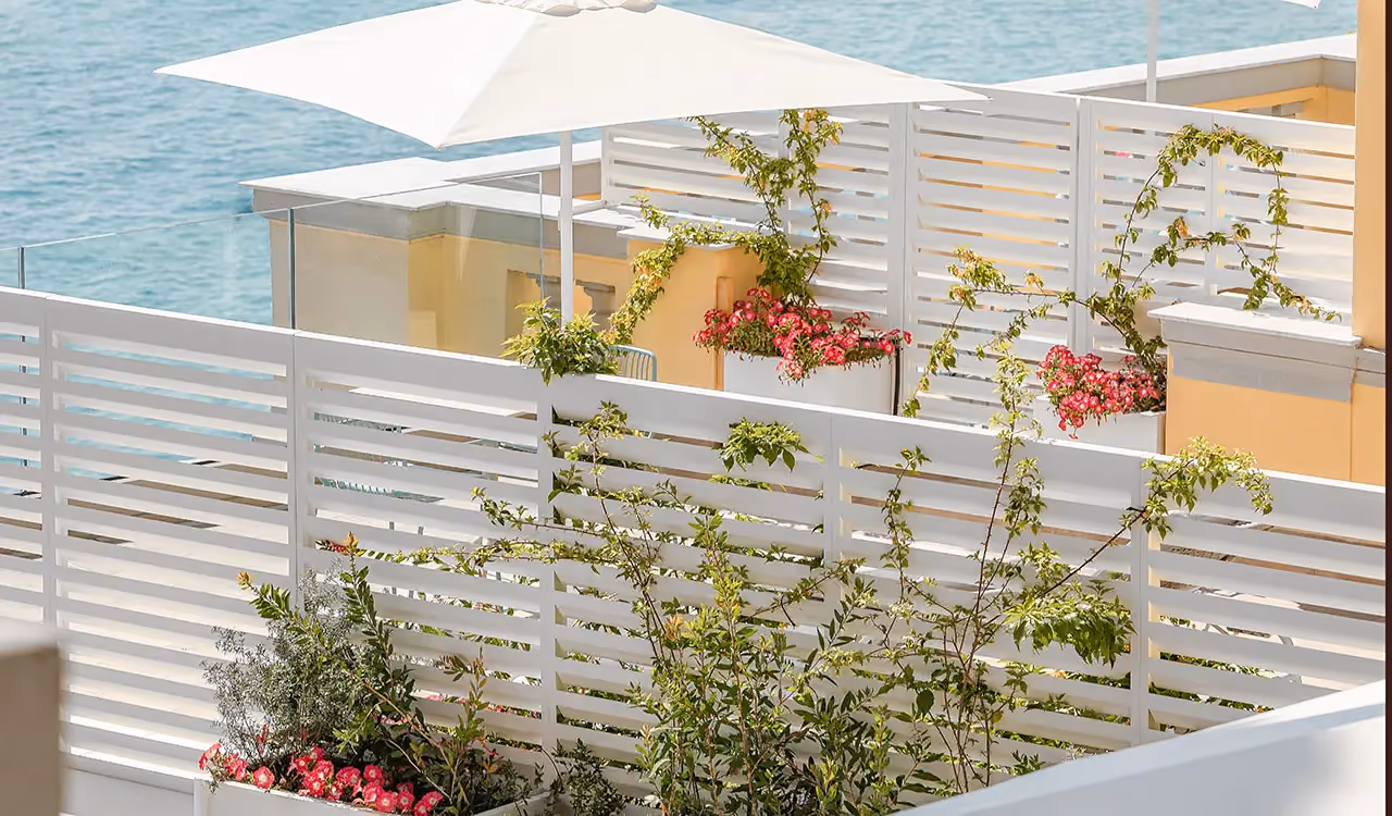 Terrasse en bord de mer avec panneaux brise-vue en bois blanc à lames, bacs rectangulaires blancs aux fleurs roses et un parasol.