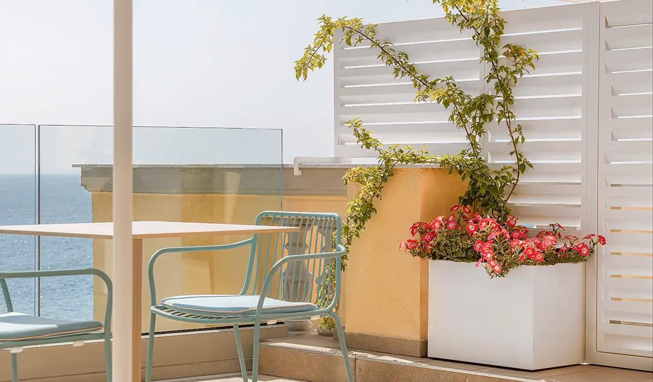 Détail d'une terrasse vue mer avec une chaise en métal bleu clair, une table basse, un bac blanc planté de bougainvillier rose et un panneau brise-vent blanc en arrière-plan.