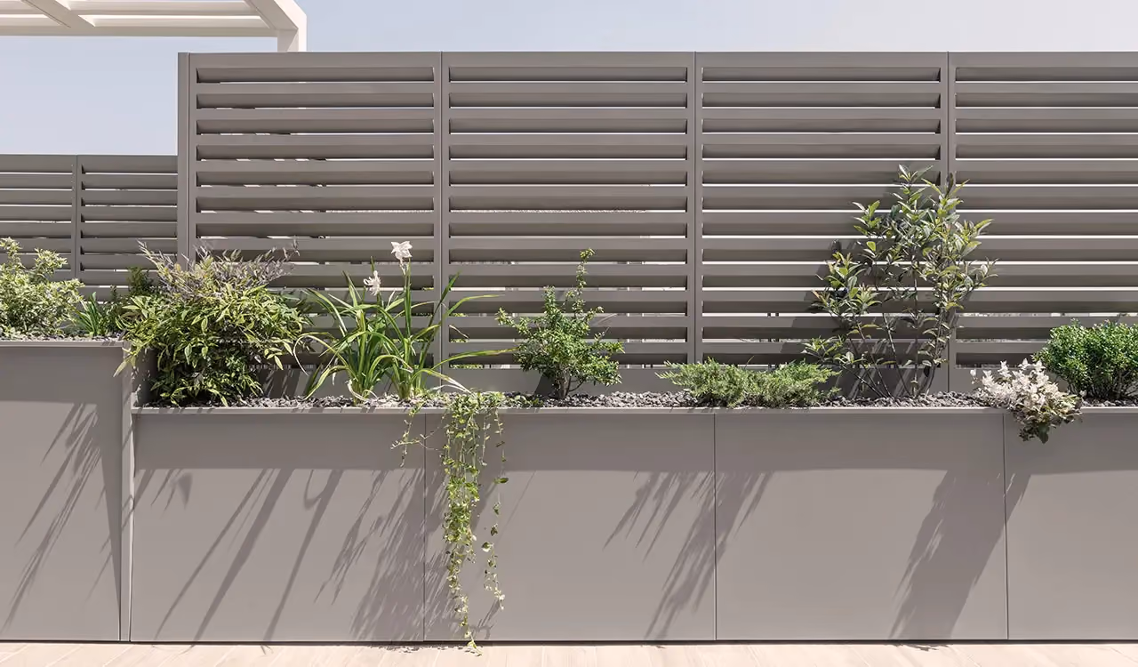 Taupe grey modular planters with green plants and flowers, set in front of a horizontally louvred windbreak panel on a modern terrace.