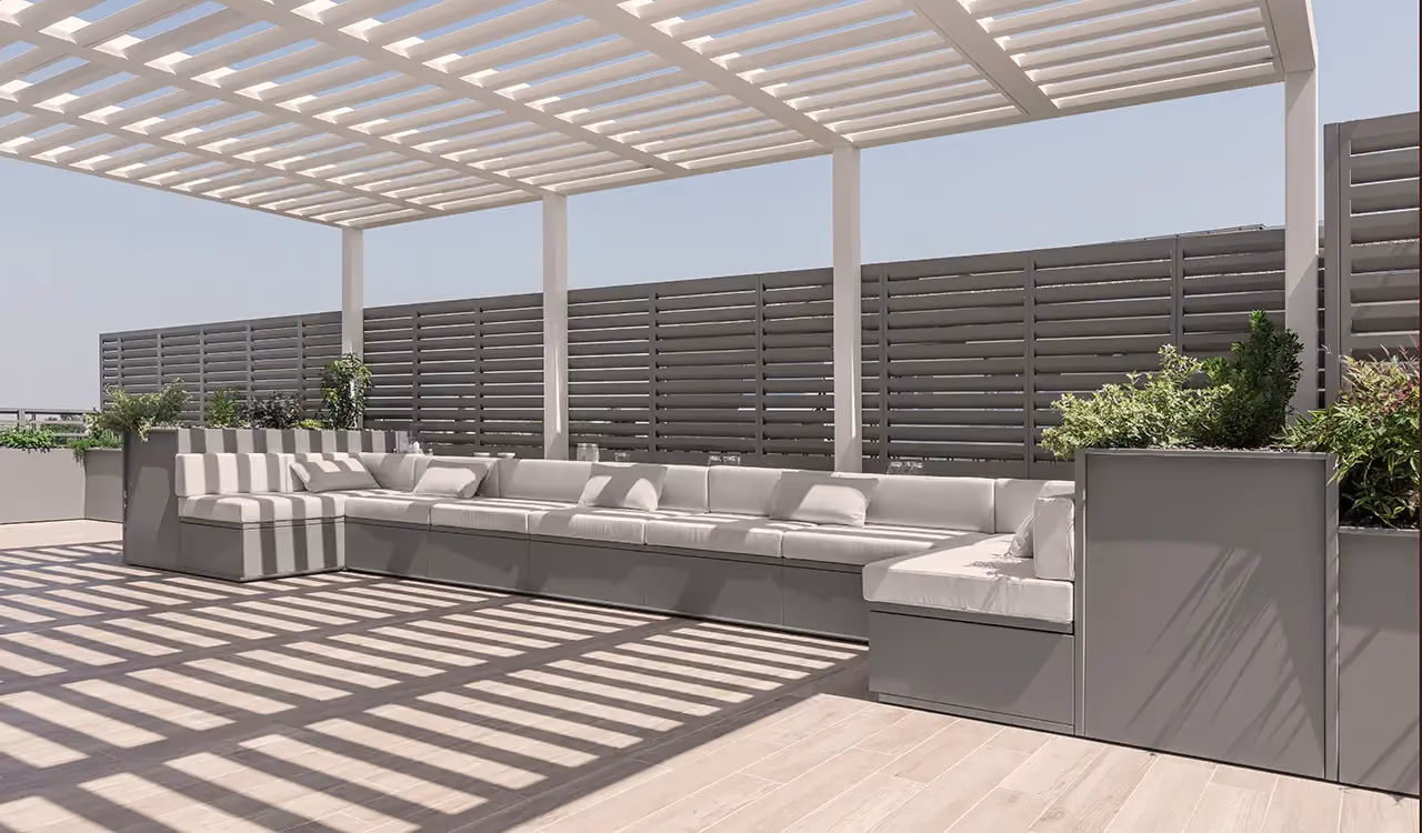 Large grey modular corner sofa with white cushions, on a terrace under a white bioclimatic pergola casting geometric shadows on the floor.