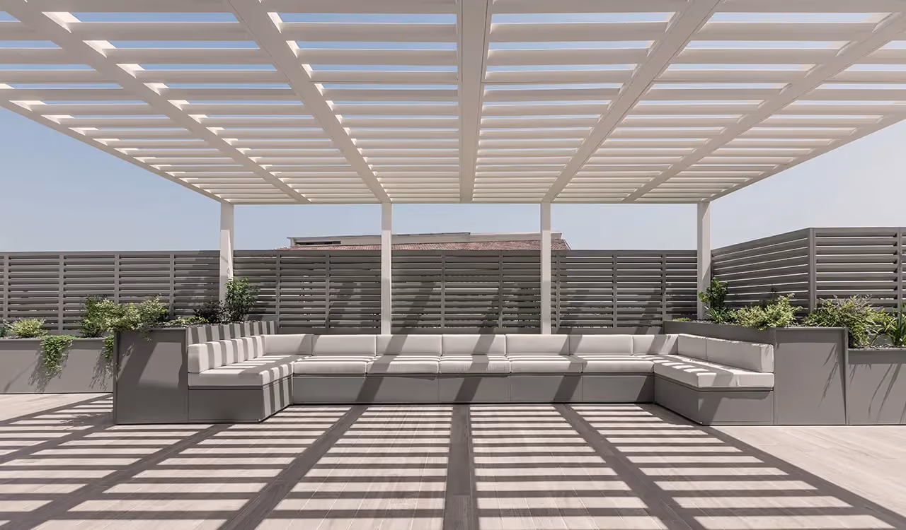 Front view of a large grey outdoor corner sofa, surrounded by planters and covered by a white louvred pergola, with windbreak panels behind.